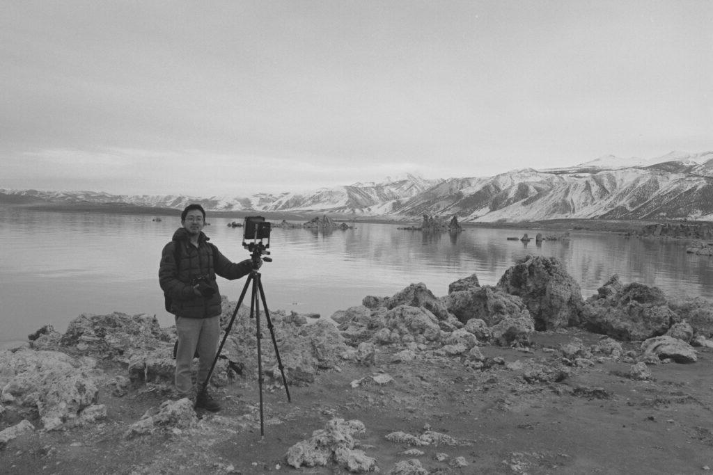 郭蓓华站在三脚架相机旁的黑白照片，在湖边，背景是山.